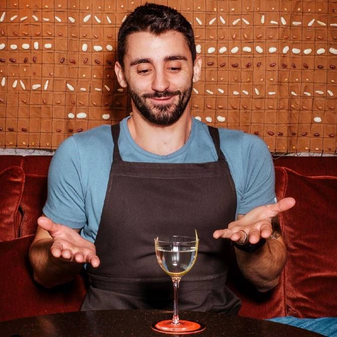 Bartender gesturing at a cocktail glass in front of a textured, patterned wall at Seed Library in London.