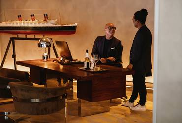 Two men in suits talking at a desk with wine glasses and a large ship model.