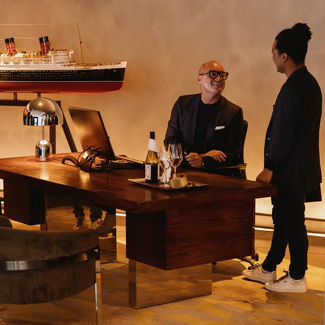 Two men in suits talking at a desk with wine glasses and a large ship model.