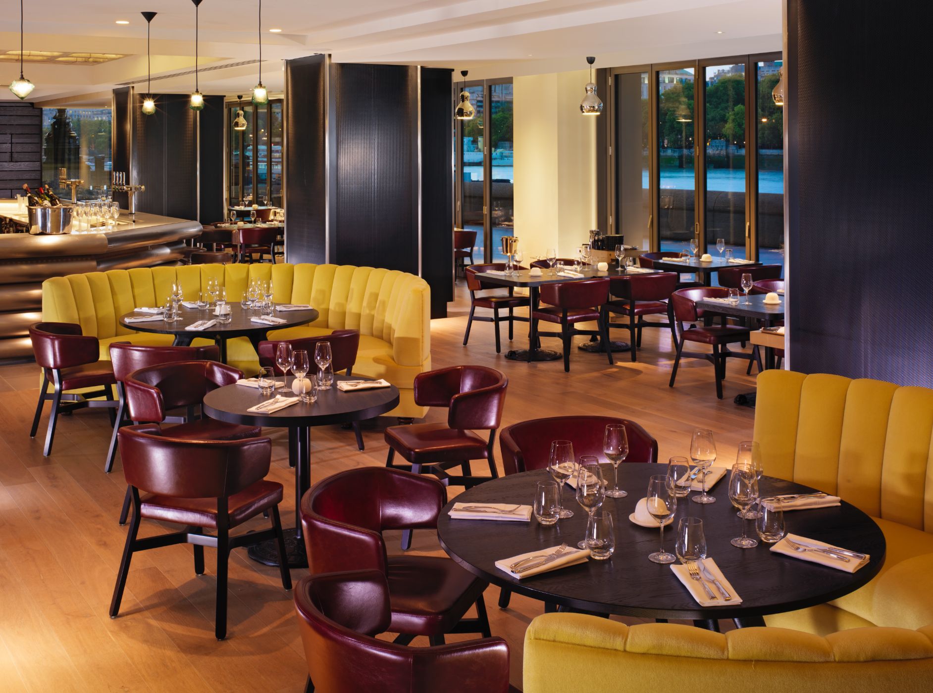 Restaurant interior featuring yellow and maroon seating, elegant table settings, and large windows overlooking the water.