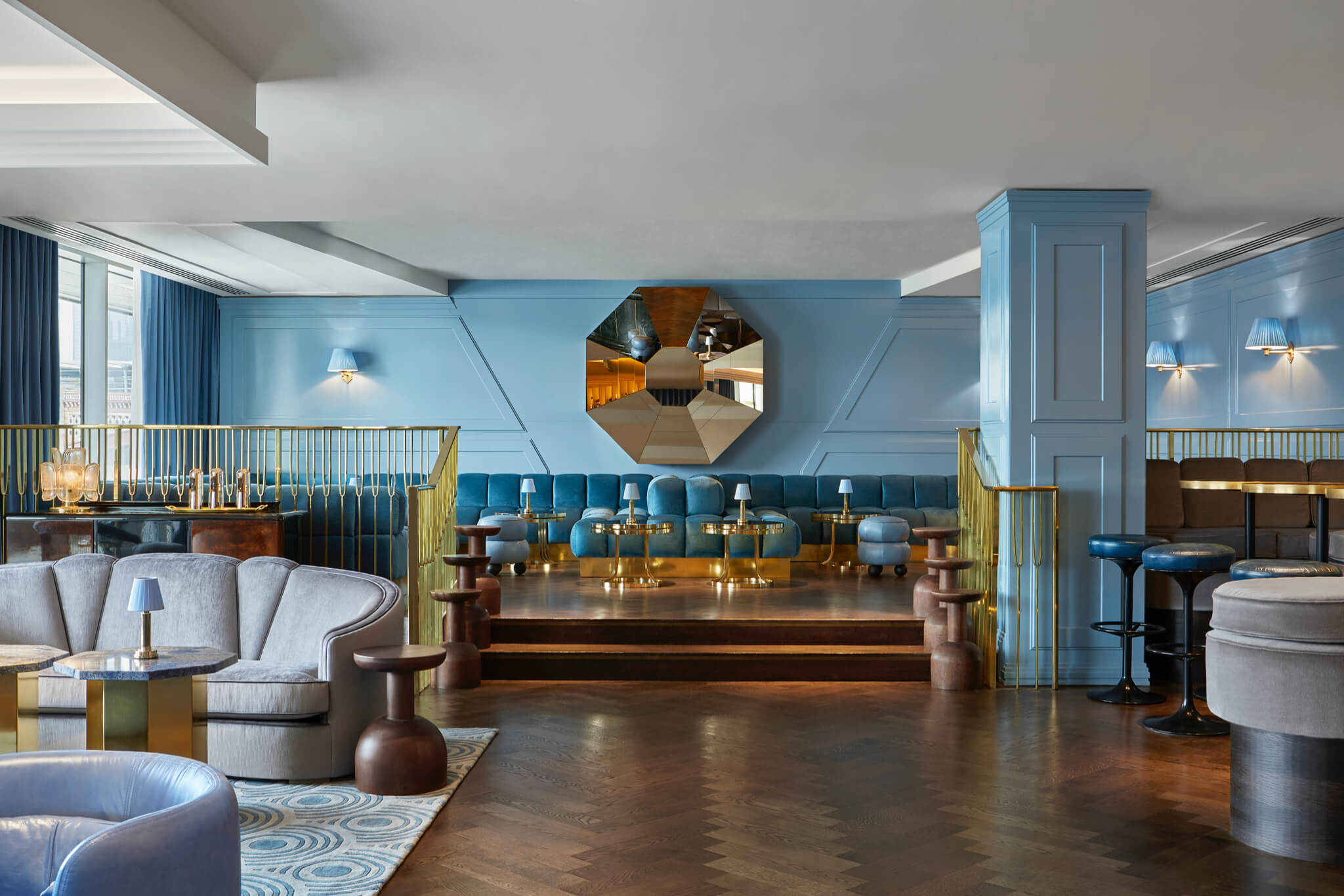 Elegant lounge area with blue upholstered seating, gold accents, and a large geometric mirror as the focal point.