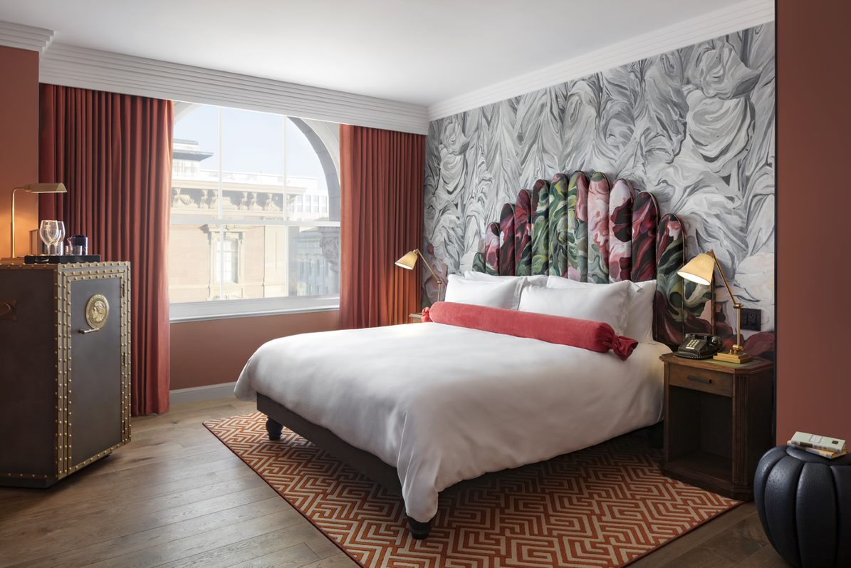 Stylish hotel bedroom at Riggs DC with floral headboard, red bolster pillow, and marbled wallpaper.