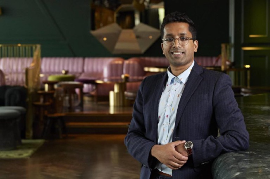 Man in a suit standing confidently in a stylish, modern lounge with purple seating and sophisticated lighting.