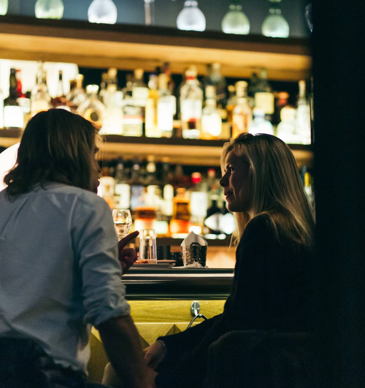 Couple talking across a bar counter with bottles lit in the background.