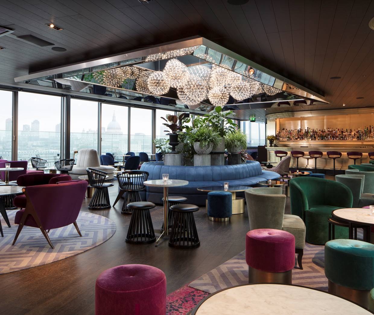 A lively rooftop bar with vibrant seating, lush greenery, and panoramic city views.