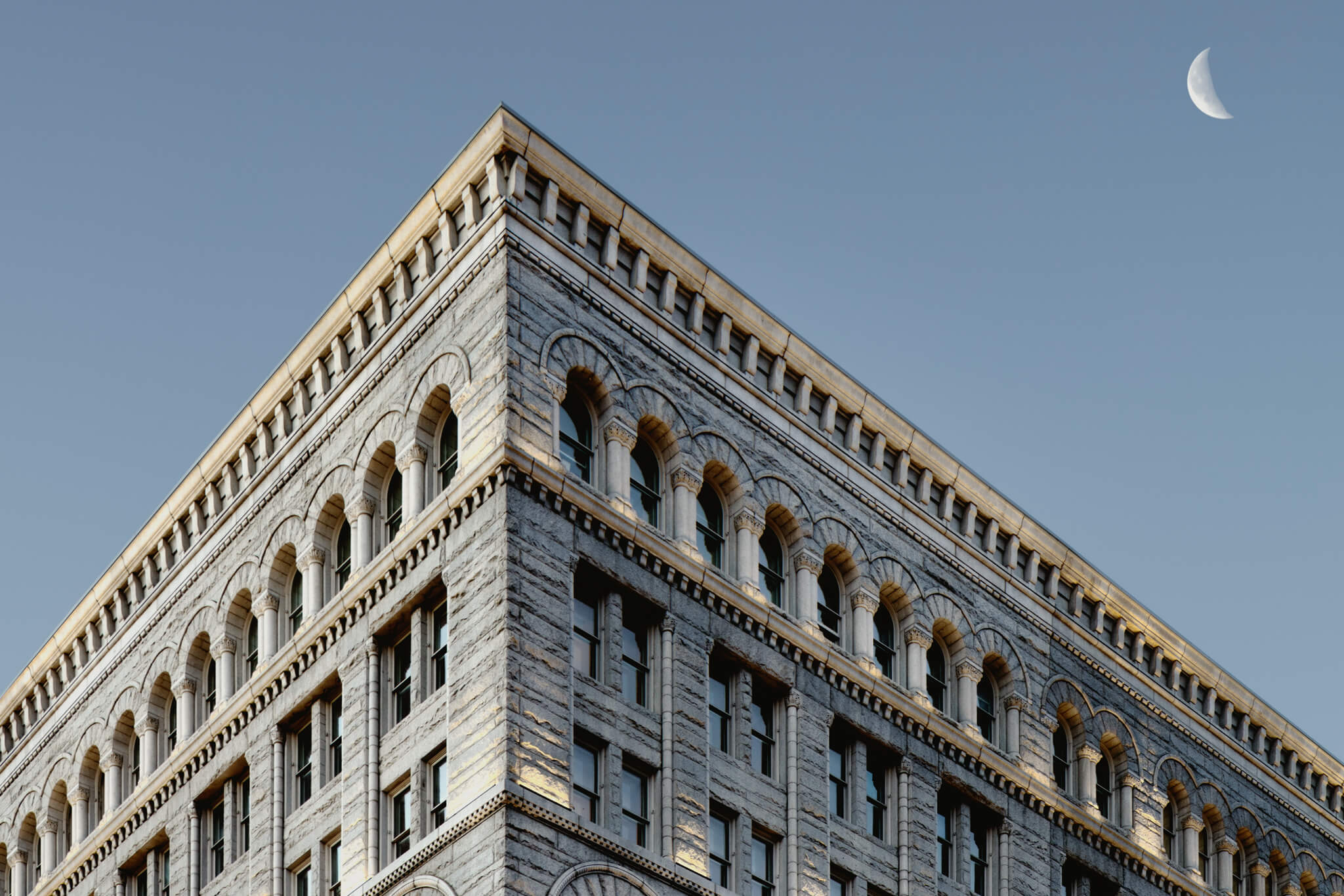 Close-up of a historic building's top corner, showcasing intricate stonework and arched windows against a clear sky.