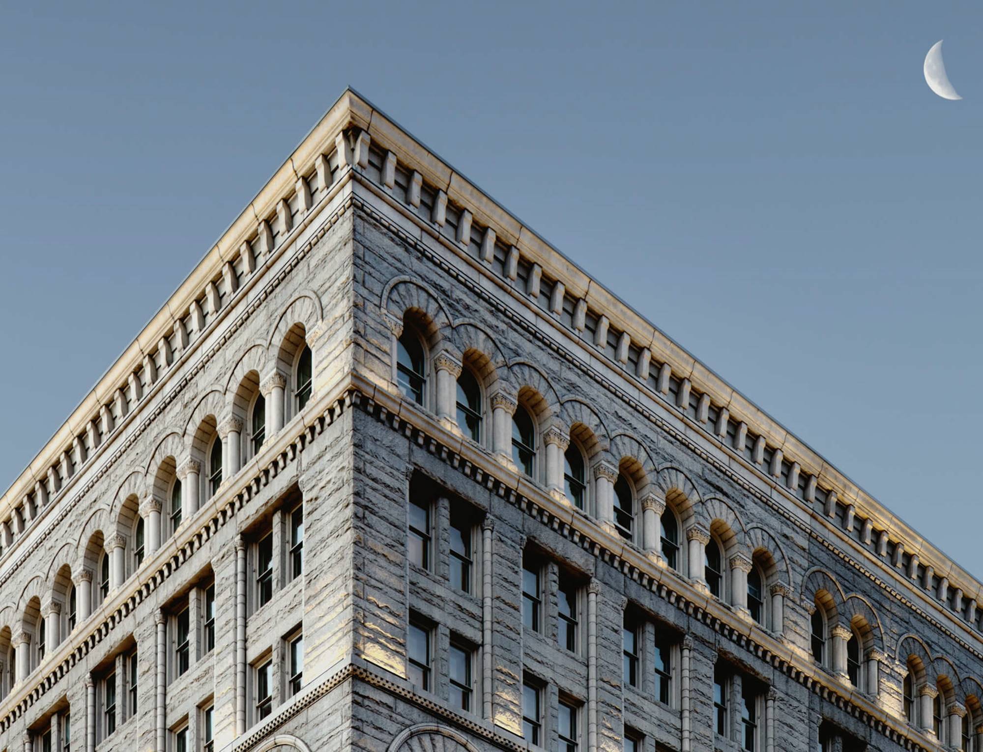 Close-up of a historic building's top corner, showcasing intricate stonework and arched windows against a clear sky.