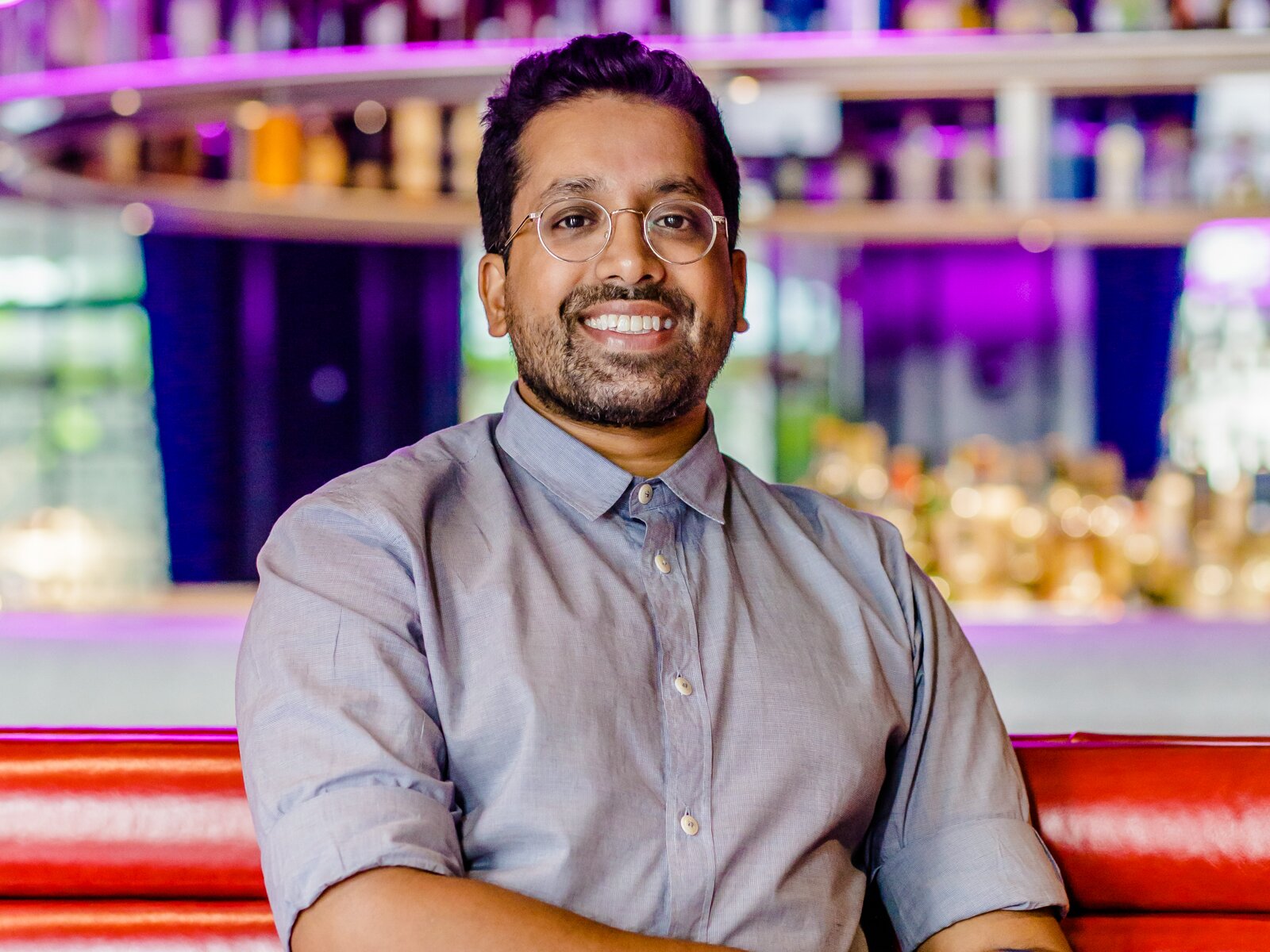Smiling man in a modern bar with purple lighting, wearing a light blue shirt, showcasing a welcoming and vibrant atmosphere.