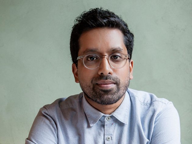 Portrait of a thoughtful man with glasses, wearing a button-up shirt, set against a muted green background.