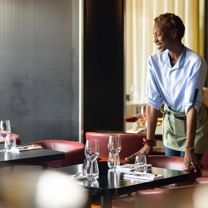 Server in blue shirt setting wine glasses at a warmly lit restaurant table.