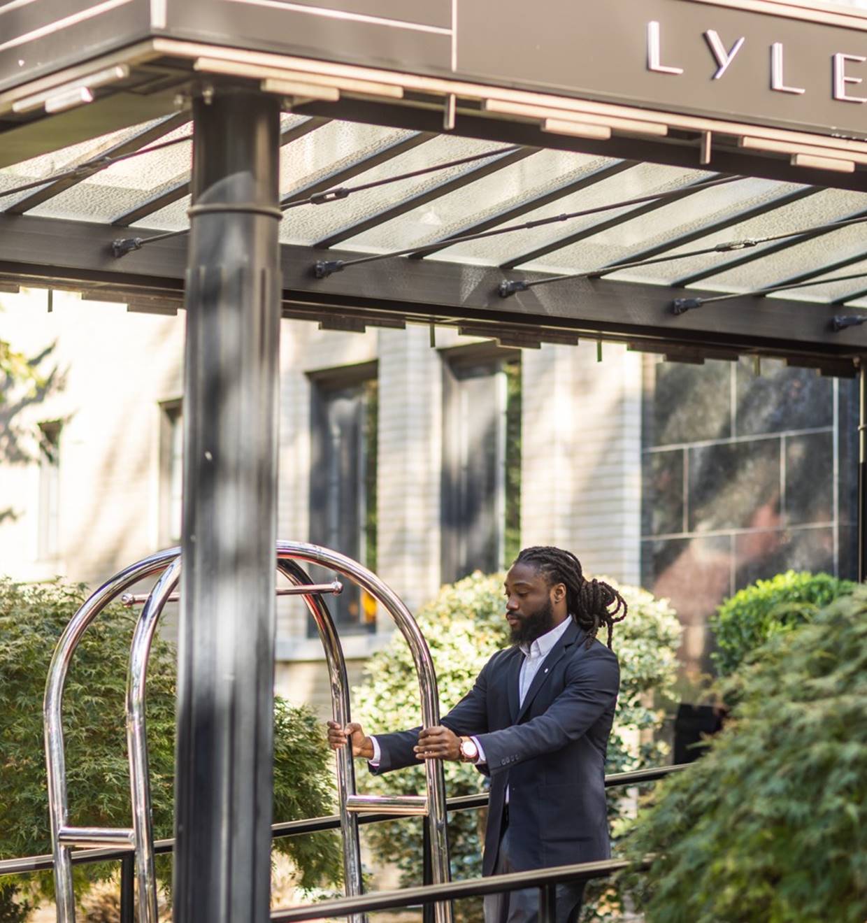 Hotel porter in a suit rolling a chrome luggage cart outside the Lyle DC hotel building under a canopy.