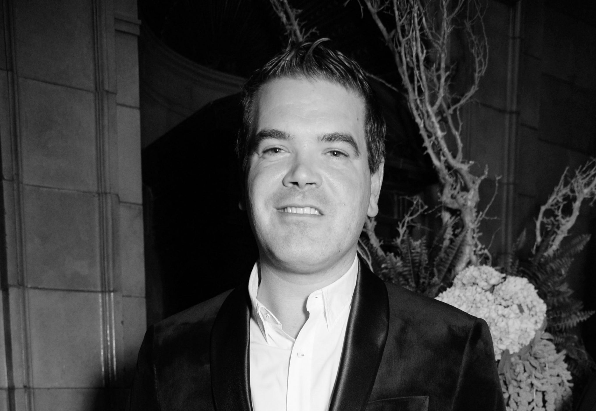 Black and white portrait of a man in formal attire, smiling at an evening event, with a sophisticated backdrop.