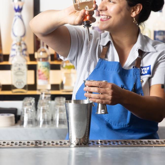 Smiling bartender in a blue apron expertly pouring liquor into a cocktail shaker at a stylish bar, showcasing mixology skills.