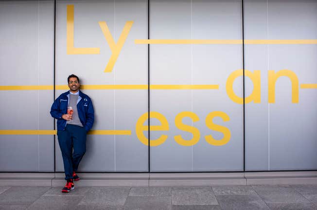 Man in a casual outfit standing against a minimalist wall with bold yellow text, holding a drink and exuding a relaxed vibe.