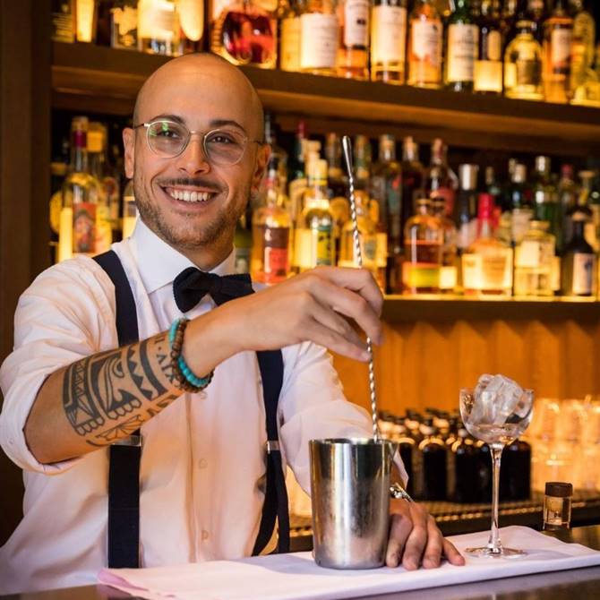 A smiling man in glasses and a tie prepares a cocktail, showcasing his mixology skills with enthusiasm and style.