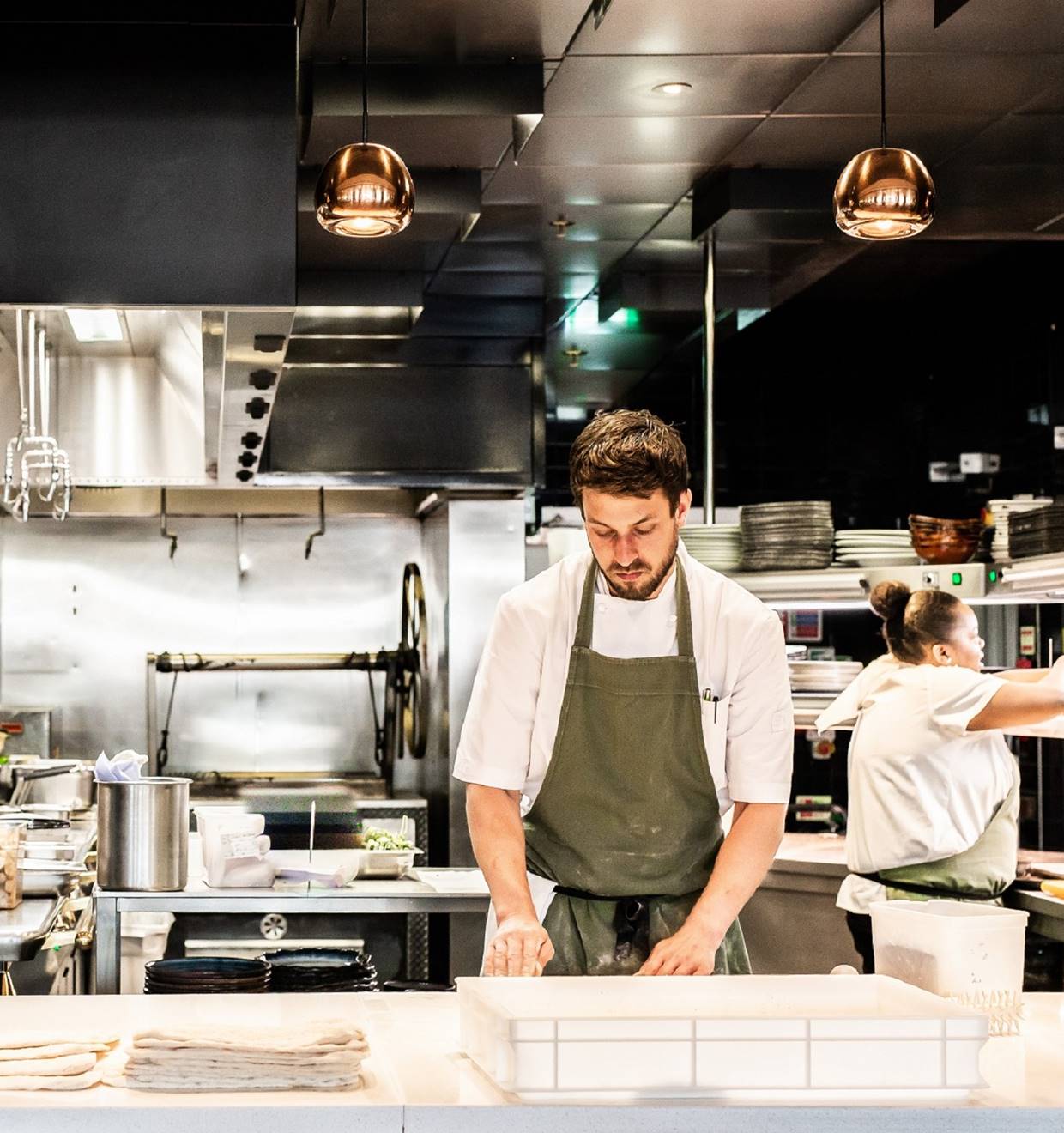 Chef in green apron preparing dough in a busy, stainless-steel kitchen.