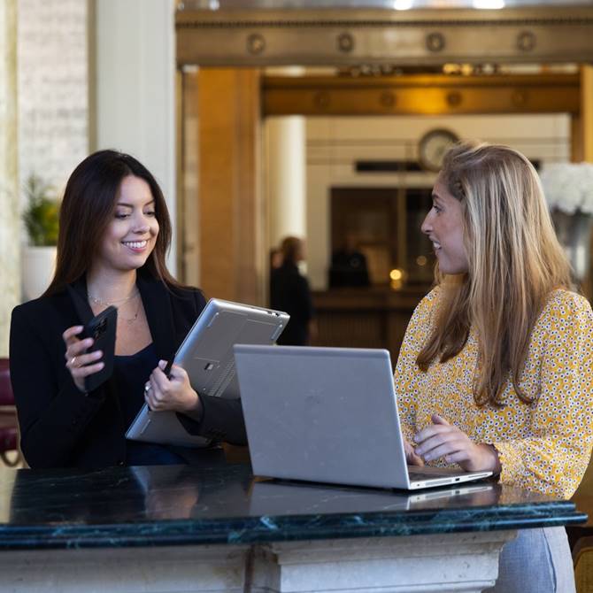 Two women engaged in work at a hotel desk, each using a laptop, focused on their tasks in a professional setting.