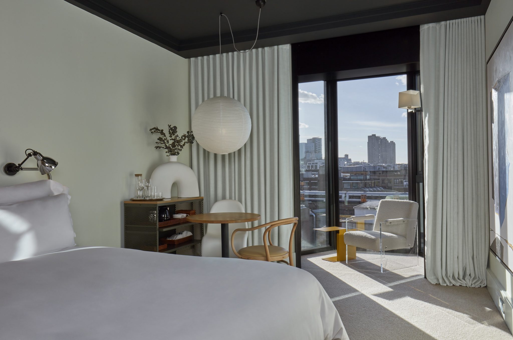 Modern hotel room with minimalist decor, featuring a large bed, floor-to-ceiling windows, and a cityscape view