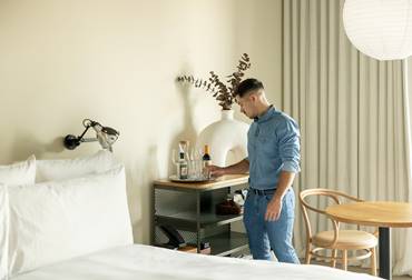Hotel staff placing wine and glasses on a minibar beside a bed in a guest room at One Hundred Shoreditch hotel.