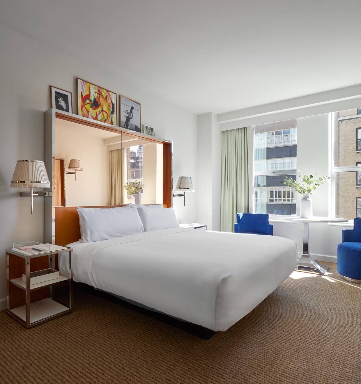 Hotel room in New York with white bedding, mirror headboard, art on wall, and city view through large window.