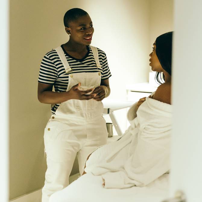 A woman in white overalls engages in conversation with another woman, both appearing attentive and engaged in discussion.