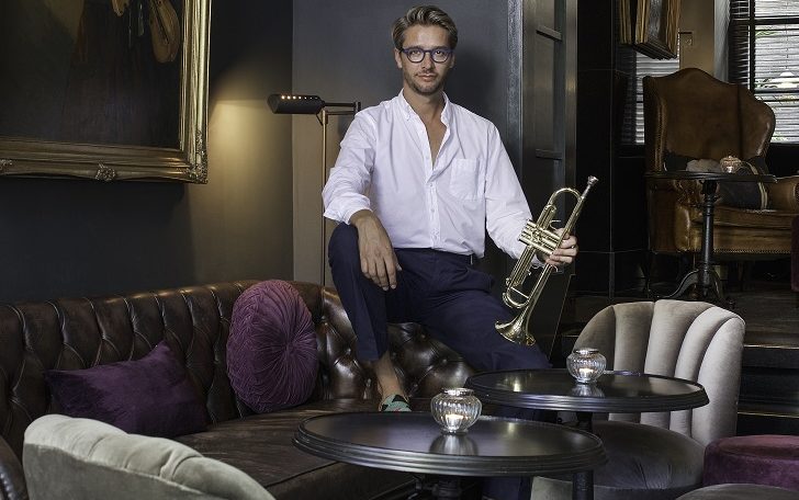 Portrait of a man in a white shirt holding a trumpet, sitting confidently on a plush, modern sofa in a stylish room.
