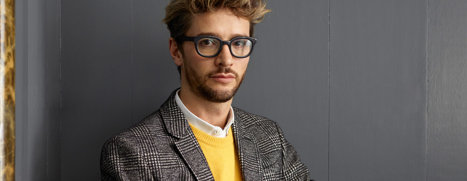 Close-up portrait of a man with glasses and a stylish outfit, set against a neutral background, conveying a modern vibe.