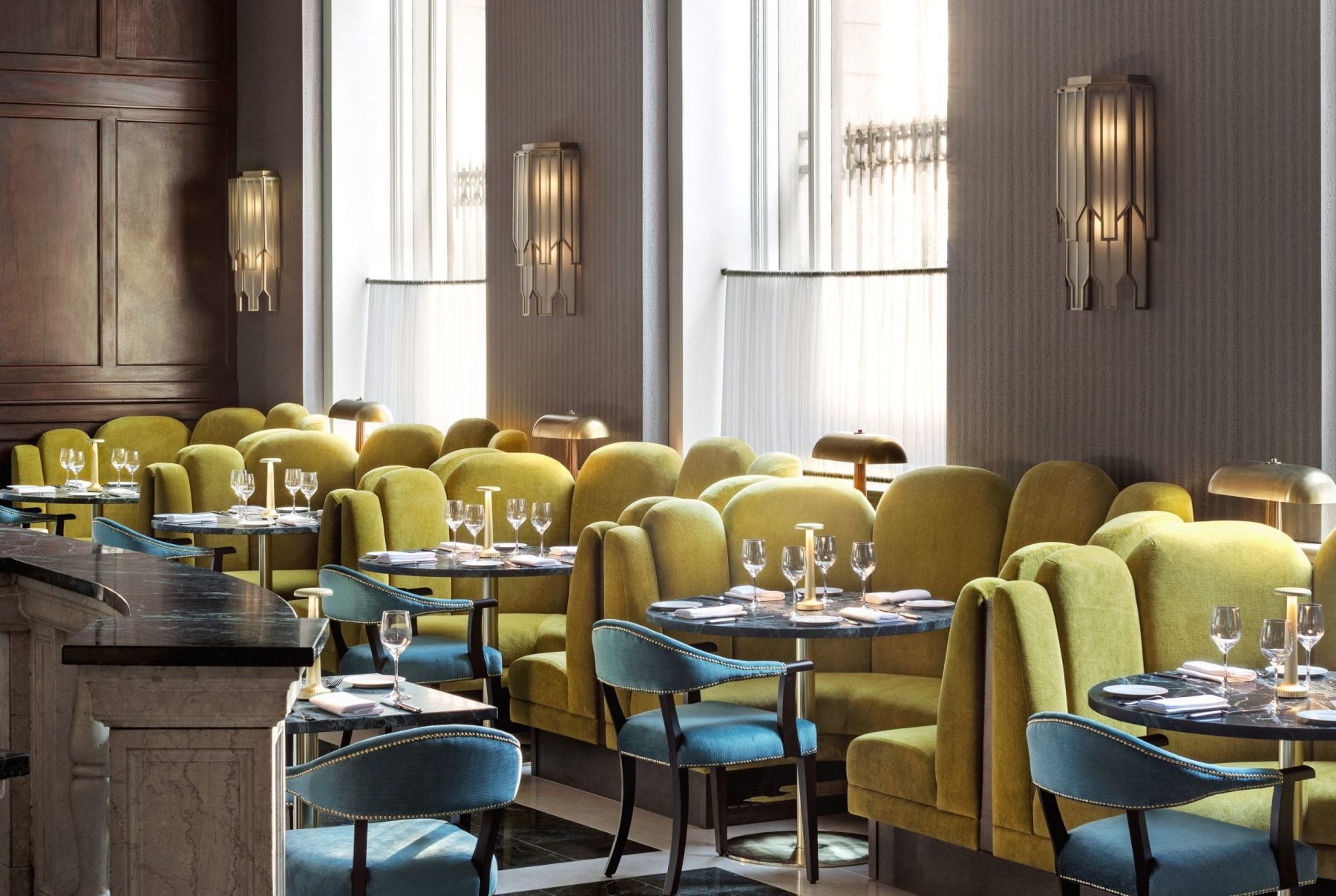 Elegant restaurant interior featuring green and blue upholstered seating, set against large windows with natural light.