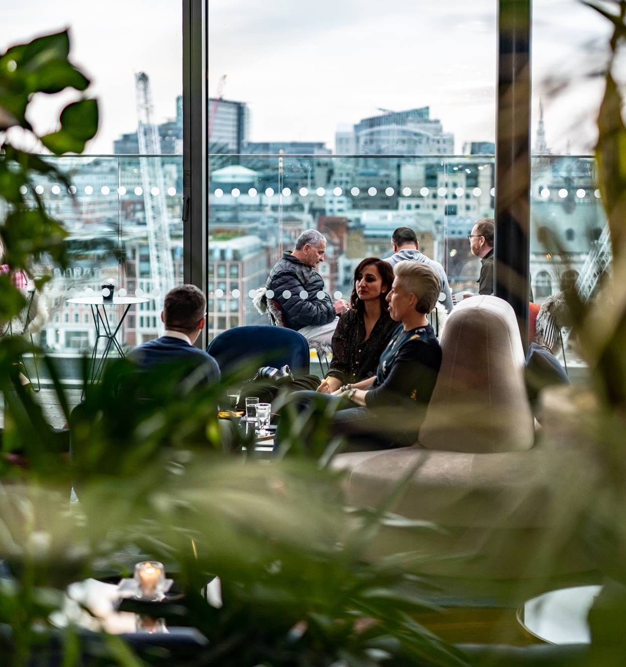 People enjoying a rooftop lounge with panoramic city views, surrounded by lush greenery and modern seating.
